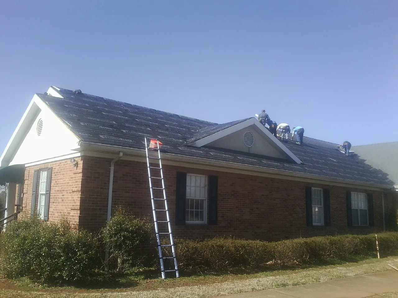 Crew tearing shingles off a brick ranch roof with a ladder set — Emergency Roof Repair work by our crew in Hunker