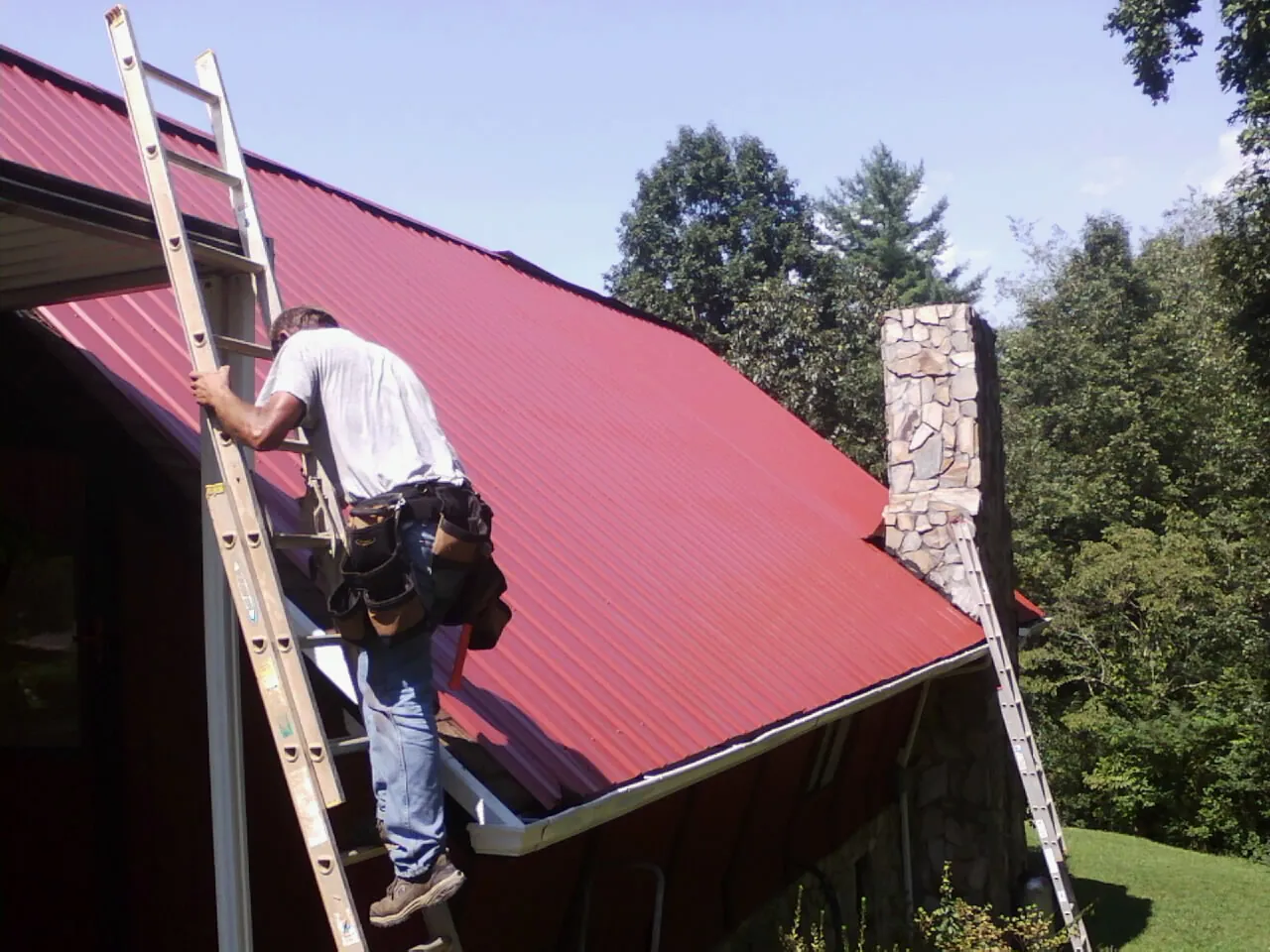 Roofer on a ladder beside a red metal roof and stone chimney — Soffit & Fascia Repair by Roofing Brothers in Hunker